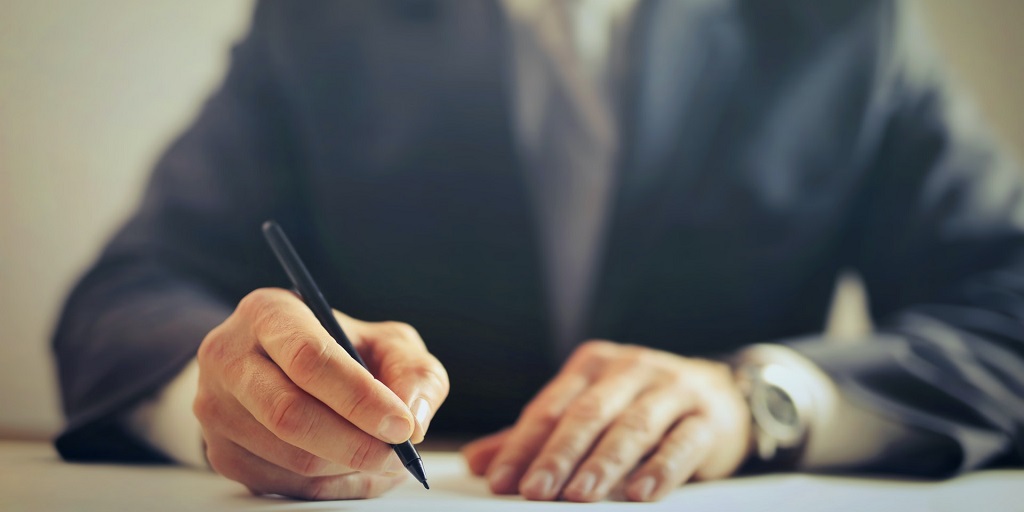 Person in a suit holding a pen, with their forearms resting on a flat white surface