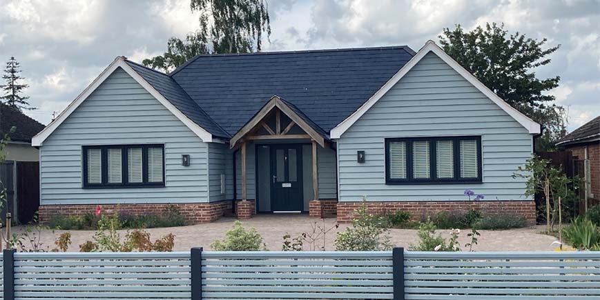 Eggshell blue cladded bungalow with wooden columned, tiled, canopy over front door and brick weave driveway surrounded by plants and flowers