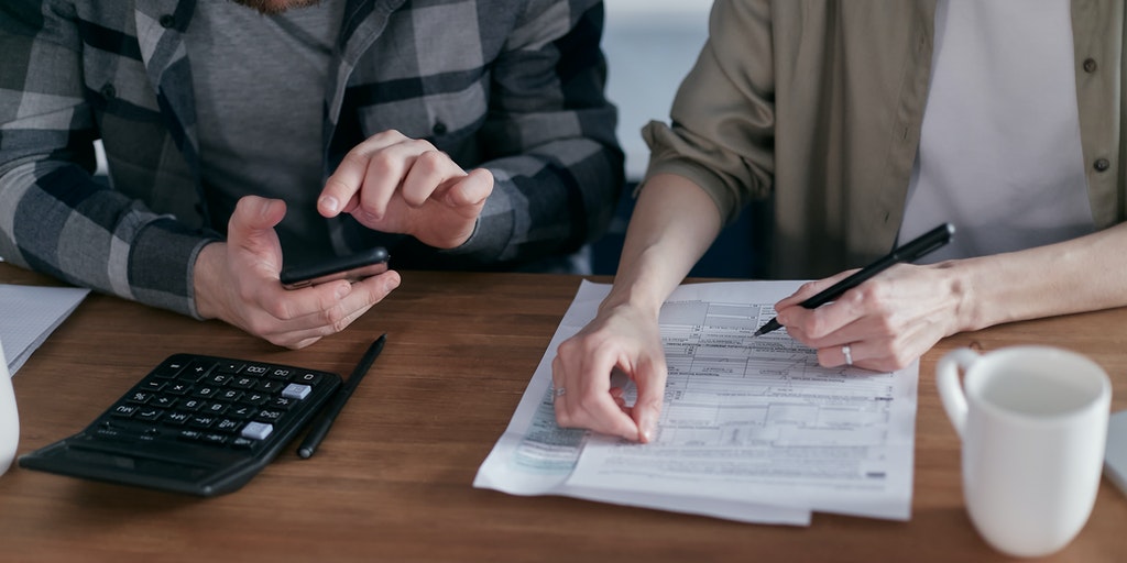 Two people sat at a wooden desk, one person holding a pen over some paperwork. There is also a calculator, pen and mug on the desk