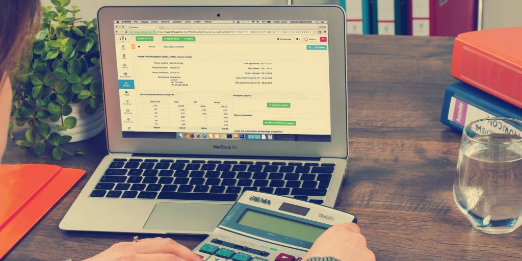 Person sat at a desk looking at a laptop and holding a calculator. There is a glass of water, plants and a folder on the desk