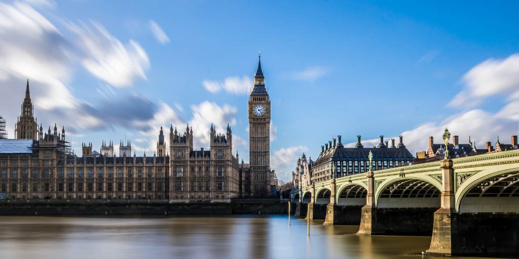 Houses of Parliament on bright sunny day with clouds in motion in the sky and Thames river in foreground