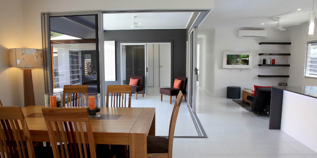 Modern, open plan apartment with dining table to left and granite kitchen counter to right, with seating and TV beyond and open courtyard accessed via sliding doors in background