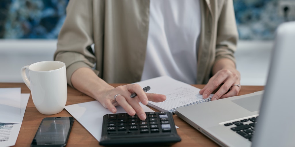 Person holding a pen, sitting at a desk with a laptop, calculator, mug, mobile phone, and papers and a notepad on it