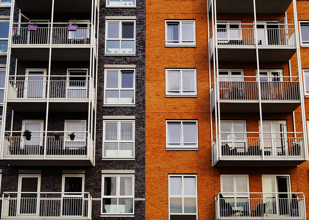 Four floors of two blocks of flats joined together, one red brick, one black bricks, both have balconies, 8 in total are visible