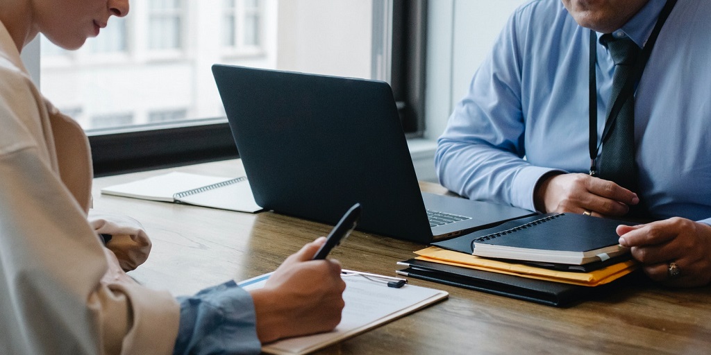 Two people facing one another across a desk. One person has a laptop infront of them, and a notebook and files, the other person is holding a pen with their hands resting on a clip board with papers on it